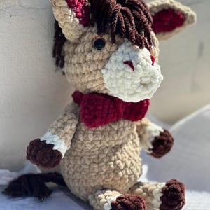 Può includere: Un peluche di asino all'uncinetto fatto a mano. L'asino è principalmente beige con dettagli marroni e bianchi. Ha un papillon, orecchie e dettagli interni delle orecchie rosso scuro. La criniera dell'asino è marrone scuro e ha un muso bianco.