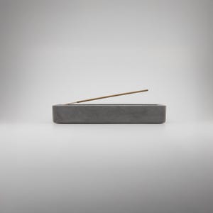 以下が含まれることがあります： 長方形の灰色のコンクリート製お香立て。お香スティックが1本あります。ホルダーはシンプルでミニマリストなデザインで、家の装飾に最適です。お香スティックは明るい茶色で、ホルダーの灰色と対照的です。