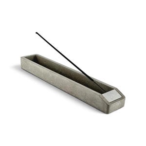 Peut inclure: Un porte-encens rectangulaire en béton gris à la surface texturée, doté d'un insert métallique argenté lisse à une extrémité. Un bâton d'encens noir fin est placé en biais, reposant dans le canal allongé du support.
