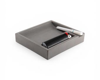 May include: A gray square ashtray with a built-in compartment. A black lighter and a white cigarette are inside the ashtray. The ashtray is made of a solid, smooth material. The items are set against a white background.