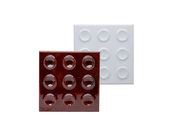 May include: Two square tiles, one brown and one white, each with nine raised, circular bumps. The brown tile has a glossy finish, while the white tile has a matte finish. The tiles are likely decorative or functional.