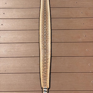 May include: A long, narrow, preserved rattlesnake skin featuring a repeating diamond pattern in shades of tan, brown, and cream. The skin is bordered by a dark, stitched edge and terminates in a distinct black and white banded tail section.