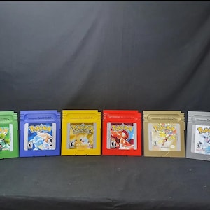 May include: A collection of six Game Boy cartridges in various colors, including green, blue, yellow, red, gold, and gray. Each cartridge features the "Pokemon" logo and character illustrations. The cartridges are arranged in a row against a dark background.