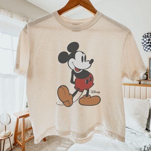 May include: Ivory-coloured t-shirt featuring a classic Mickey Mouse graphic. The cartoon character is depicted in black, red, and brown, with the Disney logo below. The shirt is hanging on a wooden hanger.