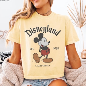 May include: Butter yellow t-shirt featuring the Disneyland logo with Mickey Mouse. The graphic includes the text "est. 1955" and "CALIFORNIA". The shirt has short sleeves and a classic crew neck.