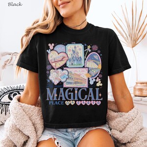 May include: Black t-shirt featuring a colorful graphic design with the text "The most MAGICAL PLACE ON EARTH." The design includes illustrations of a castle, a monorail, and other theme park elements. The shirt is a classic crew neck.