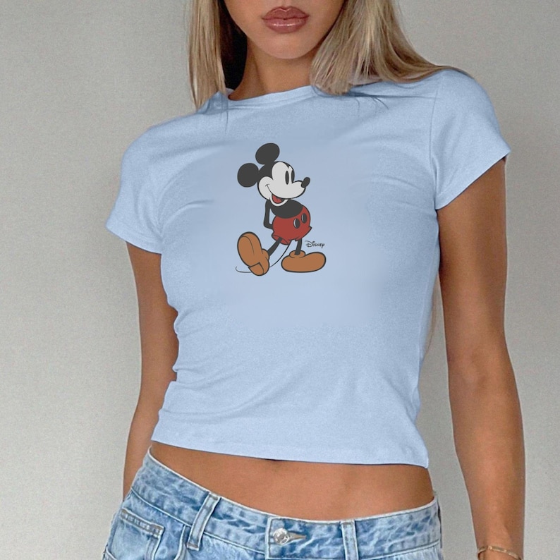 May include: Pale blue t-shirt with a Mickey Mouse graphic. The cartoon character is shown in his classic pose, wearing red shorts, brown shoes, and white gloves. The Disney logo is visible. The shirt has short sleeves and a crew neck.