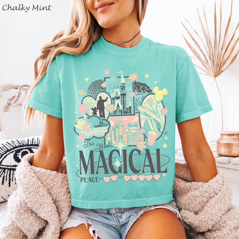 May include: A mint green short-sleeve t-shirt featuring a graphic print with various theme park icons, including a castle, a monorail, and a silhouette of a person with a mouse. The text reads The Most MAGICAL PLACE ON EARTH.