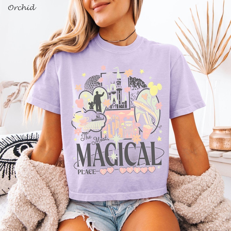 May include: A light purple short-sleeve t-shirt featuring a graphic print with the text The Most MAGICAL PLACE ON EARTH. The design includes illustrations of a castle, a statue, stars, hearts, and park icons in shades of yellow, pink, and white.