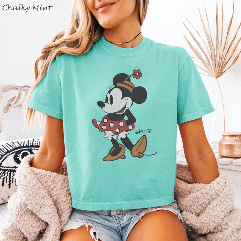 以下が含まれることがあります： ミントグリーンのTシャツ。ミニーマウスのグラフィックがプリントされています。ミニーは赤い水玉模様のスカート、茶色の靴、花のついた帽子を着用しています。「Disney」の文字がキャラクターの下に印刷されています。