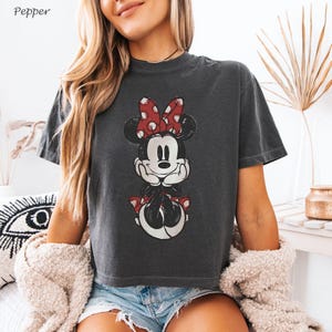 K&ouml;nnte beinhalten: Dunkelgraues T-Shirt mit einem verblassten Minnie Mouse-Motiv und einer roten Schleife mit Punkten. Das Shirt hat einen Rundhalsausschnitt und kurze &Auml;rmel. Das Wort "Pepper" befindet sich in der oberen linken Ecke.
