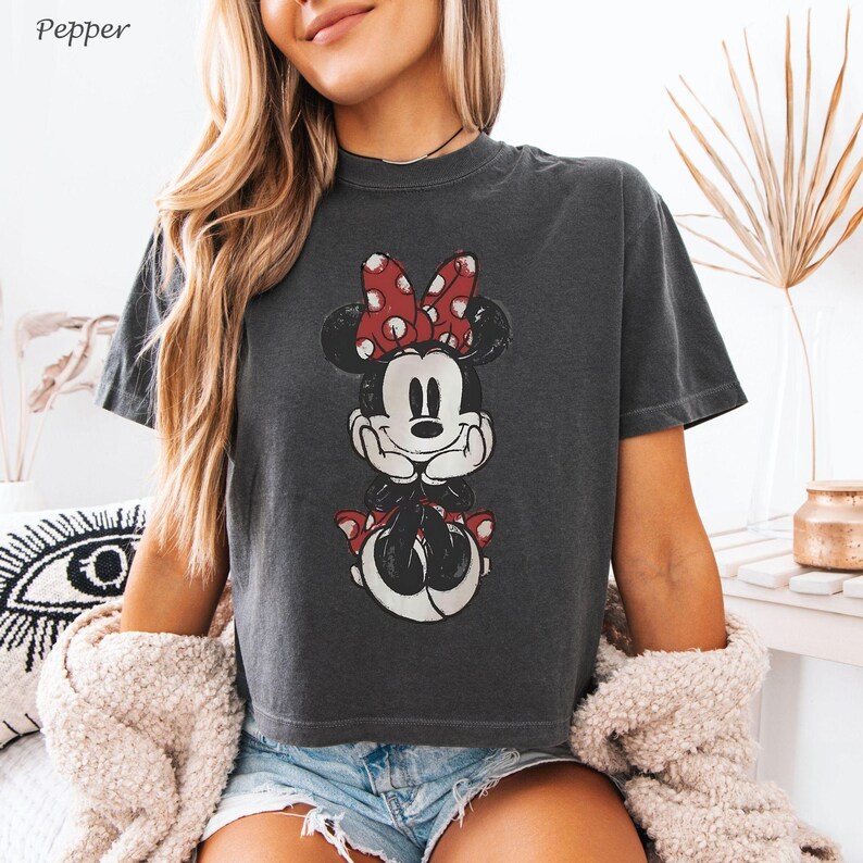 K&ouml;nnte beinhalten: Dunkelgraues T-Shirt mit einem verblassten Minnie Mouse-Motiv und einer roten Schleife mit Punkten. Das Shirt hat einen Rundhalsausschnitt und kurze &Auml;rmel. Das Wort "Pepper" befindet sich in der oberen linken Ecke.