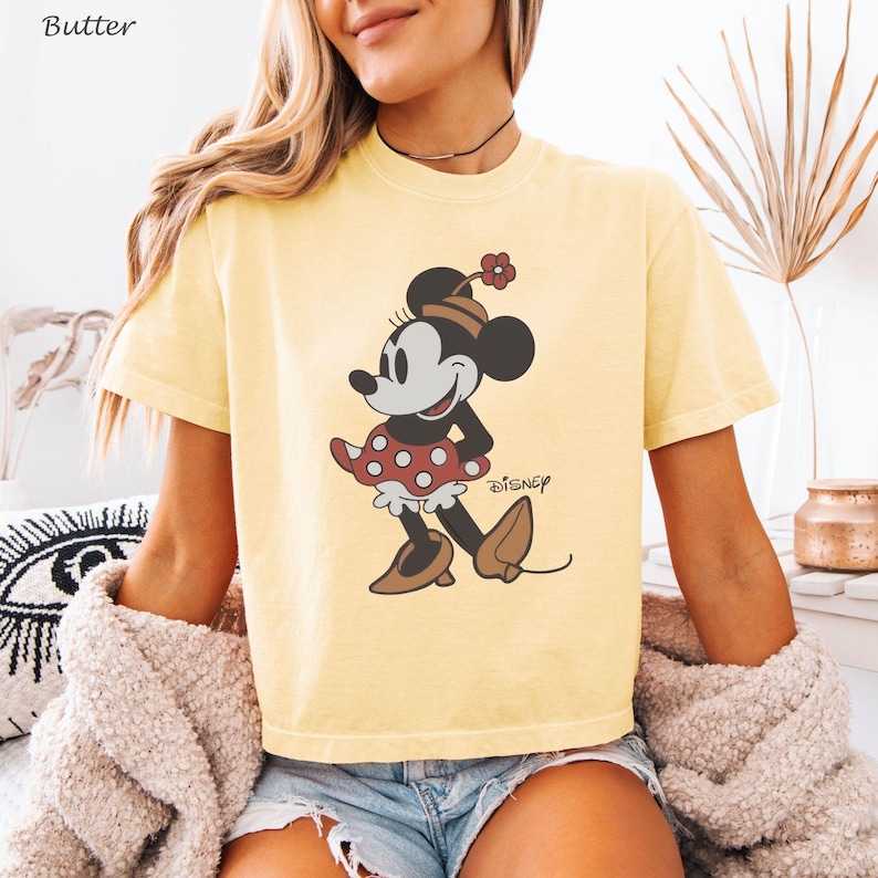 以下が含まれることがあります： バター色のTシャツ。ミニーマウスのグラフィックが特徴です。漫画のキャラクターは、赤い水玉模様のスカート、茶色の靴、花の帽子を身に着けています。ディズニーのロゴがキャラクターの下に印刷されています。