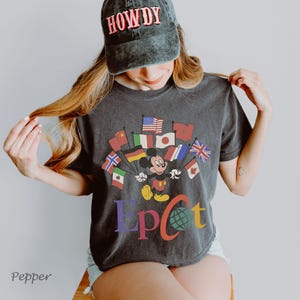 Puede incluir: Camiseta gris oscuro con Mickey Mouse sosteniendo banderas de varios pa&iacute;ses, con la palabra "EPCOT" en letras coloridas. Una gorra negra con la palabra "HOWDY" en rojo se usa en la cabeza.
