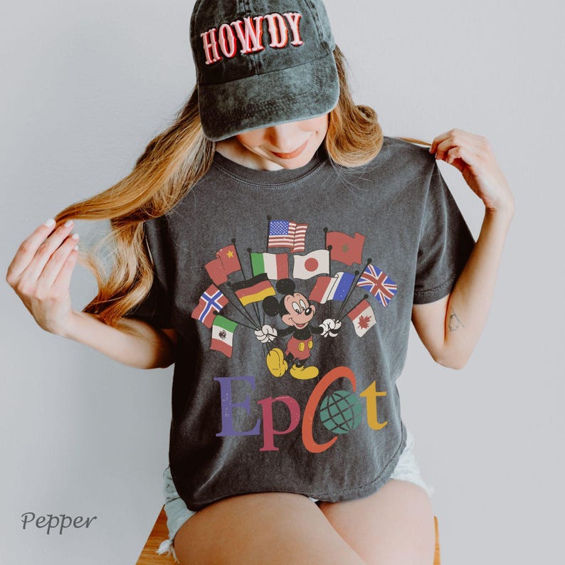 Puede incluir: Camiseta gris oscuro con Mickey Mouse sosteniendo banderas de varios pa&iacute;ses, con la palabra "EPCOT" en letras coloridas. Una gorra negra con la palabra "HOWDY" en rojo se usa en la cabeza.