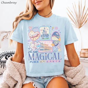 May include: Light blue t-shirt with a colorful graphic design featuring various illustrations and the text "The most MAGICAL PLACE ON EARTH." The design includes images of a castle, train, and other whimsical elements.