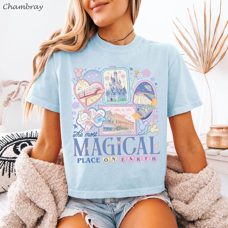 May include: Light blue t-shirt with a colorful graphic design featuring various illustrations and the text "The most MAGICAL PLACE ON EARTH." The design includes images of a castle, train, and other whimsical elements.