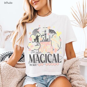 May include: A white short-sleeve t-shirt featuring a graphic print with a castle, a silhouette of a person, and various shapes. The text reads The Most MAGICAL PLACE ON EARTH in black and pink lettering, accented with yellow and pink flowers.