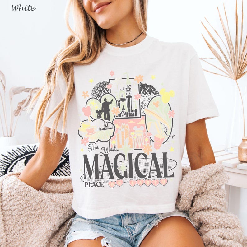 May include: A white short-sleeve t-shirt featuring a graphic print with a castle, a silhouette of a person, and various shapes. The text reads The Most MAGICAL PLACE ON EARTH in black and pink lettering, accented with yellow and pink flowers.