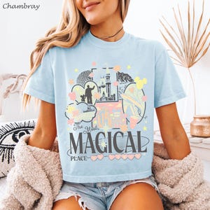 May include: A light blue t-shirt featuring a graphic print of a castle, a monorail, and various park icons. The text reads The Most MAGICAL PLACE ON EARTH. The design includes pastel-colored hearts, stars, and clouds in yellow, pink, and white.