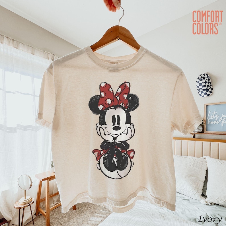 May include: Ivory-colored t-shirt featuring a graphic of Minnie Mouse with a red polka-dot bow. The Comfort Colors brand shirt is hanging on a wooden hanger. The text "Comfort Colors" is visible in the upper right corner.