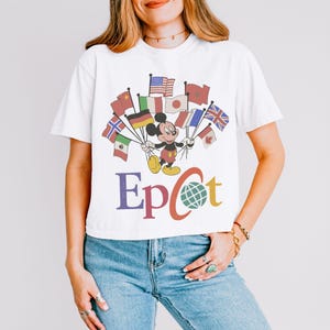 Puede incluir: Camiseta blanca con Mickey Mouse sosteniendo banderas de varios pa&iacute;ses. La palabra "Epcot" est&aacute; impresa debajo, con un globo reemplazando la letra "o". La camiseta tiene un estilo retro, con colores descoloridos.