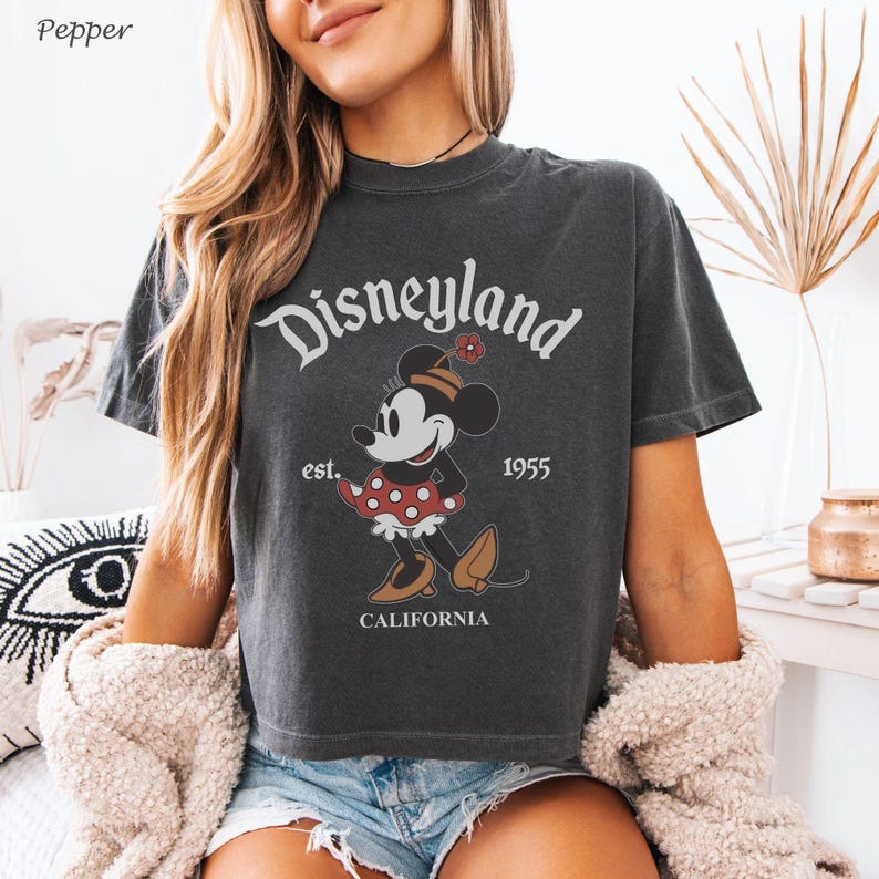 May include: Dark gray t-shirt featuring a graphic of Minnie Mouse in a red polka dot dress, with the text "Disneyland" above and "est. 1955 CALIFORNIA" below. The shirt has a vintage, worn look.