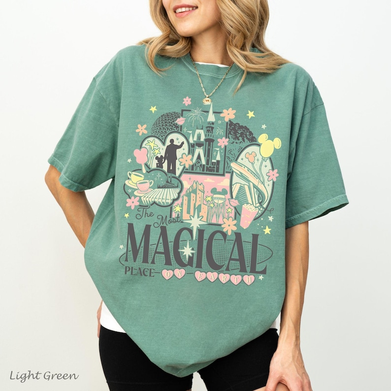 May include: A sage green short-sleeve t-shirt featuring a graphic print with the text The Most MAGICAL PLACE ON EARTH. The design includes illustrations of a castle, teacups, fireworks, and stars in shades of pink, yellow, and dark gray.