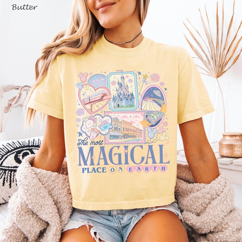 May include: Butter yellow t-shirt featuring a colorful graphic design with the text "The most MAGICAL PLACE ON EARTH." The design includes illustrations of a castle, a monorail, and other theme park elements. The shirt has short sleeves and a crew neck.