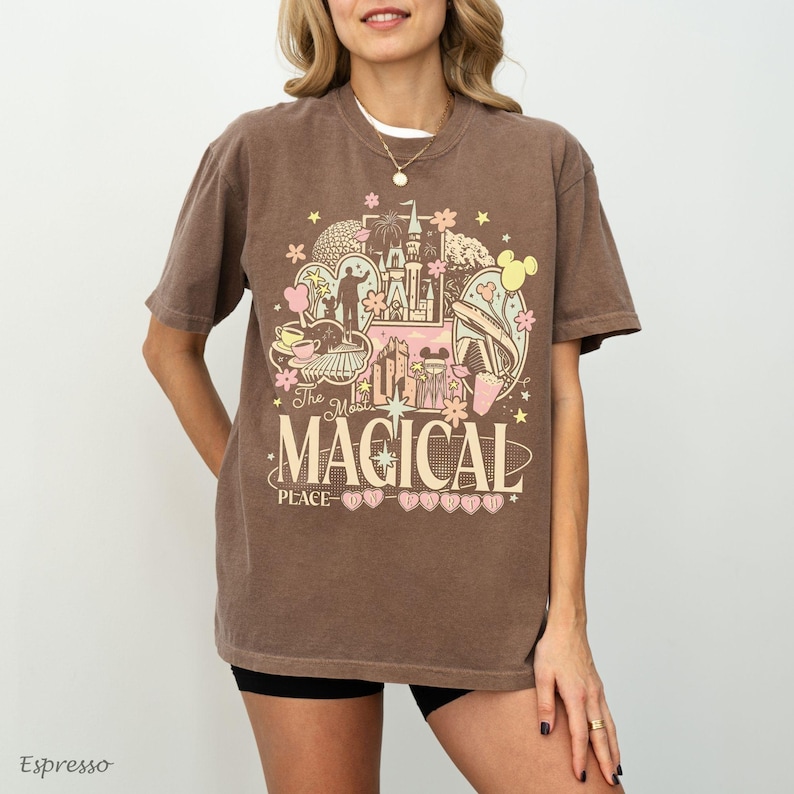 May include: A brown short-sleeve t-shirt featuring a graphic print with the text The Most MAGICAL PLACE ON EARTH. The design includes illustrations of a castle, flowers, stars, and park-themed icons in shades of pink, yellow, and white.