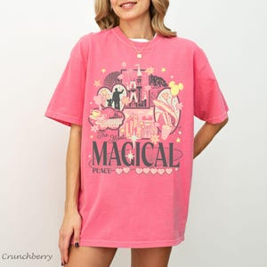 May include: A pink short-sleeve t-shirt featuring a graphic print with a castle, stars, and various shapes. The text reads The Most MAGICAL PLACE with a series of small hearts underneath. The design uses shades of yellow, white, and dark gray.