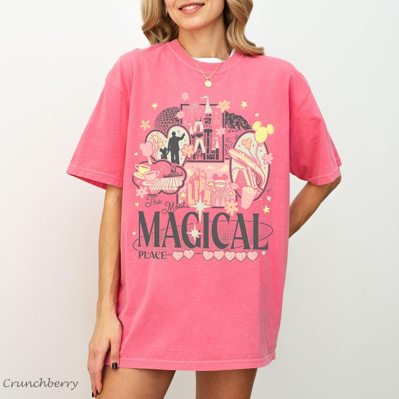 May include: A pink short-sleeve t-shirt featuring a graphic print with a castle, stars, and various shapes. The text reads The Most MAGICAL PLACE with a series of small hearts underneath. The design uses shades of yellow, white, and dark gray.