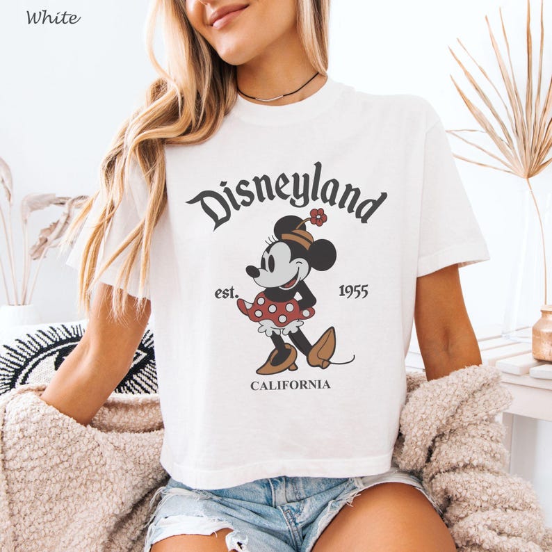 May include: White t-shirt featuring a vintage Disneyland graphic. The design includes the Disneyland logo, a Minnie Mouse illustration, and the text "est. 1955 CALIFORNIA". The shirt has short sleeves and a crew neck.