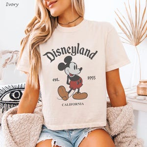 May include: Ivory-colored t-shirt featuring a vintage Disneyland graphic. The design includes Mickey Mouse, the word "Disneyland," and the text "est. 1955 CALIFORNIA." The shirt has short sleeves and a crew neck.