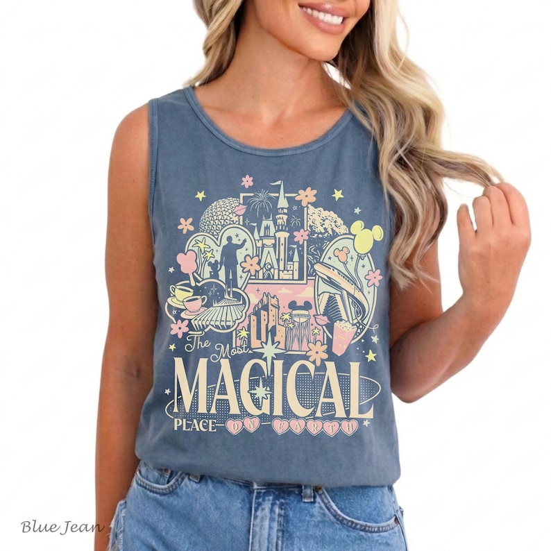 May include: A slate blue sleeveless tank top featuring a graphic print with various theme park landmarks, stars, and flowers. The text reads The Most MAGICAL PLACE ON EARTH in cream-colored lettering, accented by small pink heart shapes.