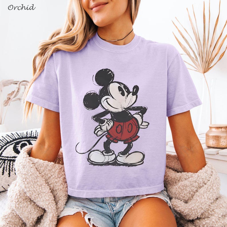 K&ouml;nnte beinhalten: Ein helllila T-Shirt mit einem Mickey Mouse-Motiv im Used-Look. Die Zeichentrickfigur ist in Schwarz, Wei&szlig; und Rot gehalten, mit den H&auml;nden in den H&uuml;ften. Das Wort "Orchid" befindet sich in der oberen linken Ecke.