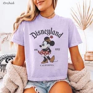 May include: A light purple t-shirt featuring the word "Disneyland" in a stylized font, along with a graphic of Minnie Mouse. The shirt also includes the text "est. 1955 CALIFORNIA".