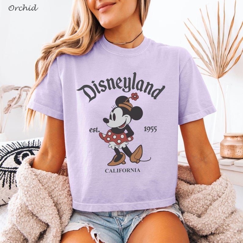 May include: A light purple t-shirt featuring the word "Disneyland" in a stylized font, along with a graphic of Minnie Mouse. The shirt also includes the text "est. 1955 CALIFORNIA".