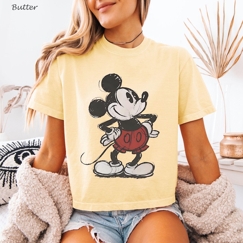 May include: Butter yellow t-shirt featuring a classic Mickey Mouse graphic. The cartoon character is depicted in black, white, and red, with a vintage, sketched style. The shirt has short sleeves and a crew neck.