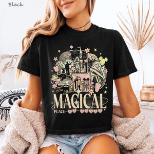 May include: A black short-sleeve t-shirt featuring a graphic print with various theme park icons, including a castle, fireworks, and a monorail. The text reads The Most MAGICAL PLACE ON EARTH in cream and pink lettering with small flower accents.
