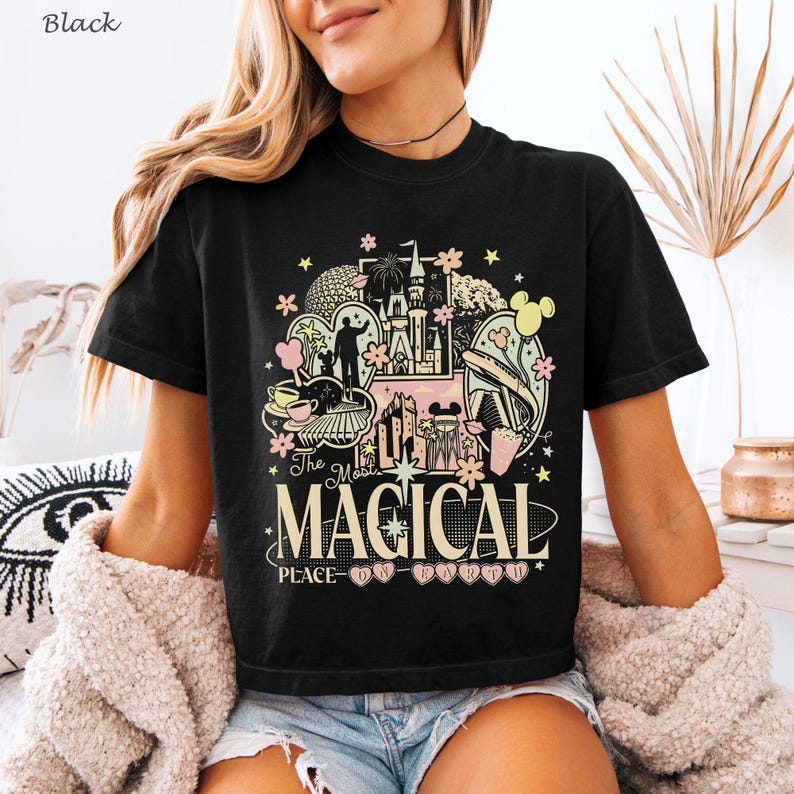 May include: A black short-sleeve t-shirt featuring a graphic print with various theme park icons, including a castle, fireworks, and a monorail. The text reads The Most MAGICAL PLACE ON EARTH in cream and pink lettering with small flower accents.