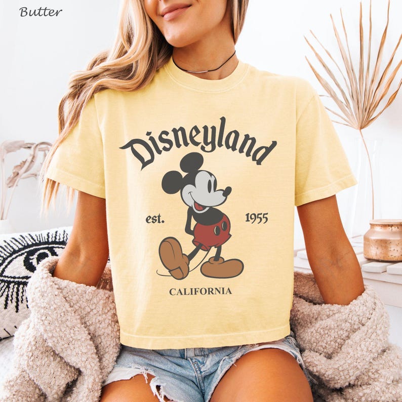 May include: Butter yellow t-shirt featuring the Disneyland logo with Mickey Mouse. The graphic includes the text "est. 1955" and "CALIFORNIA". The shirt has short sleeves and a classic crew neck.