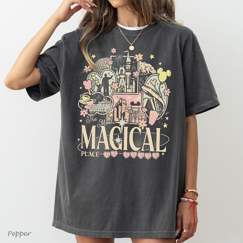 May include: A charcoal gray short-sleeve t-shirt featuring a multi-colored graphic print of a castle, fireworks, and park icons. The text reads The Most MAGICAL PLACE ON EARTH. The design includes pink, yellow, and white accents on the dark fabric.