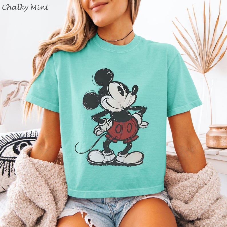 K&ouml;nnte beinhalten: T&uuml;rkisfarbenes T-Shirt mit einem verblichenen Mickey Mouse-Motiv. Die Cartoonfigur ist in Schwarz, Wei&szlig; und Rot dargestellt. Der Text "Chalky Mint" befindet sich oben links. Das Shirt hat kurze &Auml;rmel.