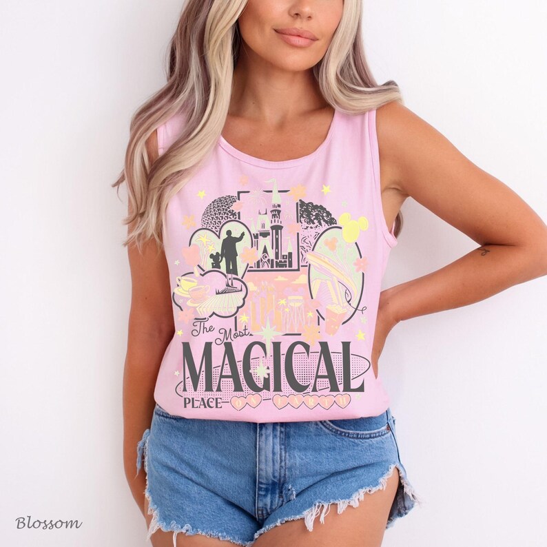 May include: A light pink sleeveless tank top featuring a graphic print of a castle, a silhouette of a person, and various icons. The text reads The Most MAGICAL PLACE in dark gray lettering, accented by small yellow stars and pastel-colored shapes.