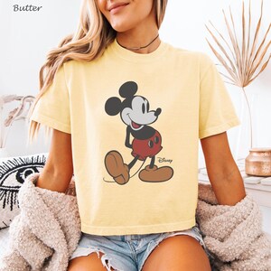 May include: A pale yellow t-shirt with a Mickey Mouse graphic. The cartoon character is in black, red, and brown, with the Disney logo. The shirt has short sleeves and a crew neck. The word "Butter" is in the top left corner.