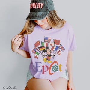 Puede incluir: Una camiseta de color lila claro con un dise&ntilde;o de Mickey Mouse sosteniendo banderas de varios pa&iacute;ses. La palabra "Epcot" est&aacute; impresa en letras coloridas debajo. La camiseta es de estilo casual.