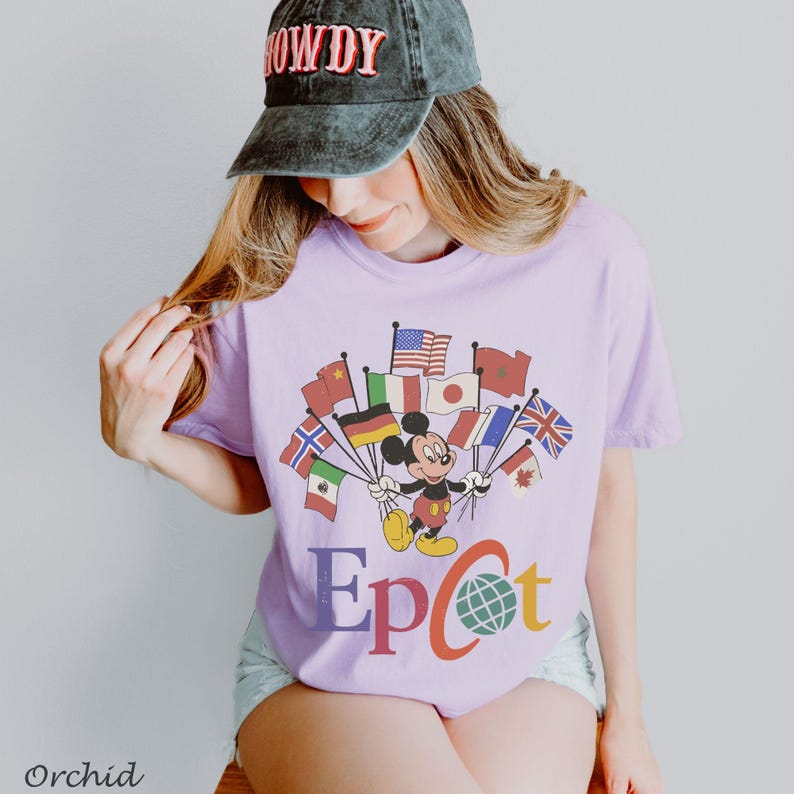 Puede incluir: Una camiseta de color lila claro con un dise&ntilde;o de Mickey Mouse sosteniendo banderas de varios pa&iacute;ses. La palabra "Epcot" est&aacute; impresa en letras coloridas debajo. La camiseta es de estilo casual.