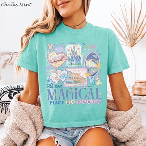 May include: A mint green t-shirt with a colorful graphic design featuring various attractions and the text "The most MAGICAL PLACE ON EARTH". The design includes images of a castle, a monorail, and other park elements.