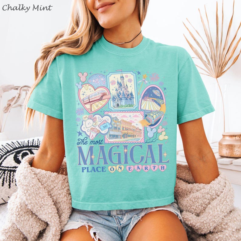 May include: A mint green t-shirt with a colorful graphic design featuring various attractions and the text "The most MAGICAL PLACE ON EARTH". The design includes images of a castle, a monorail, and other park elements.